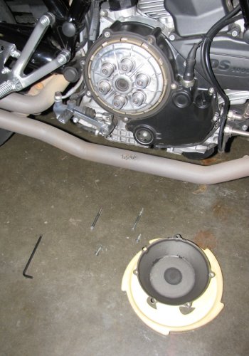 The four bolts are removed and the cover is on the ground (it's resting on a foam Frisbee).  Notice that the bolts are different lengths so make sure you put them back in the same holes.  You can also see the clutch dust in the cover and on the exhaust pipe.
